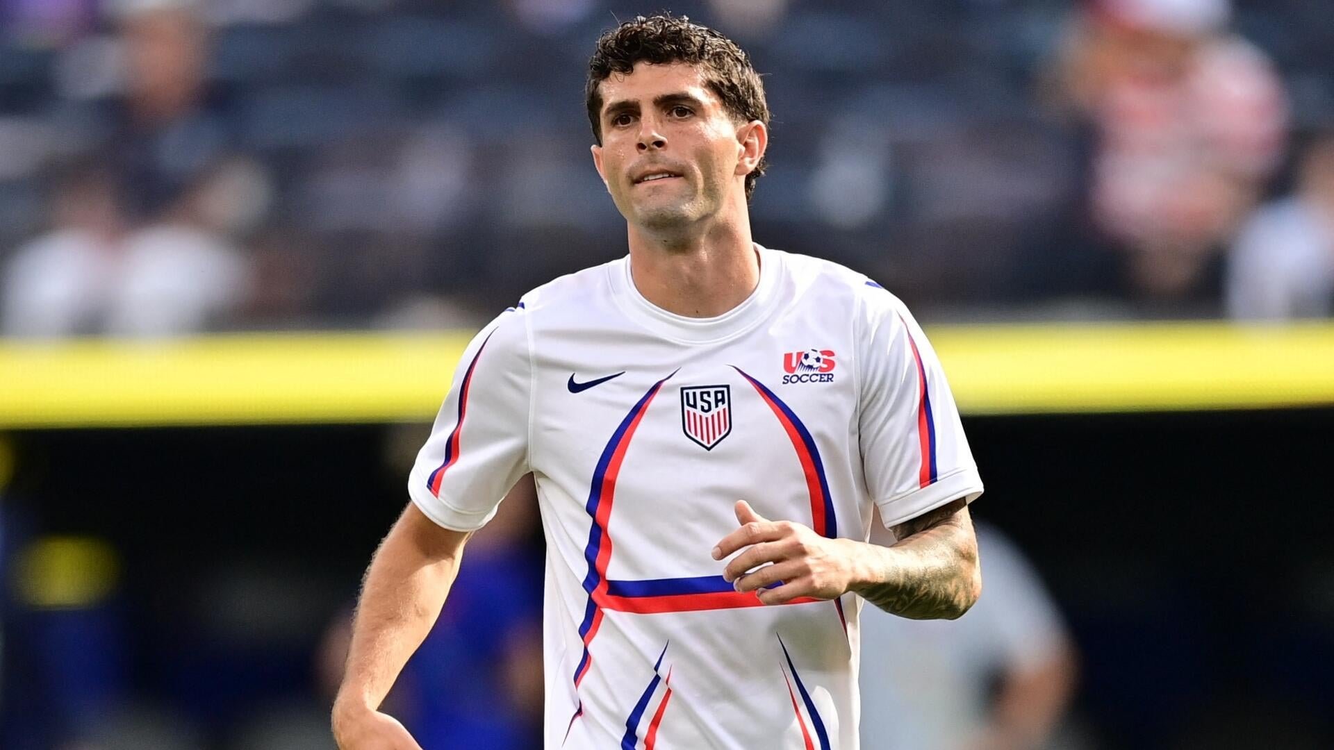 A dark-haired soccer player wearing a white USA Soccer jersey jogs past blurred spectators in a stadium setting.
