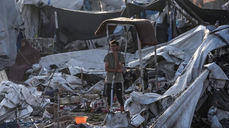 A bearded man stands amid a sprawling encampment of torn white tents and scattered debris, holding a long pole.