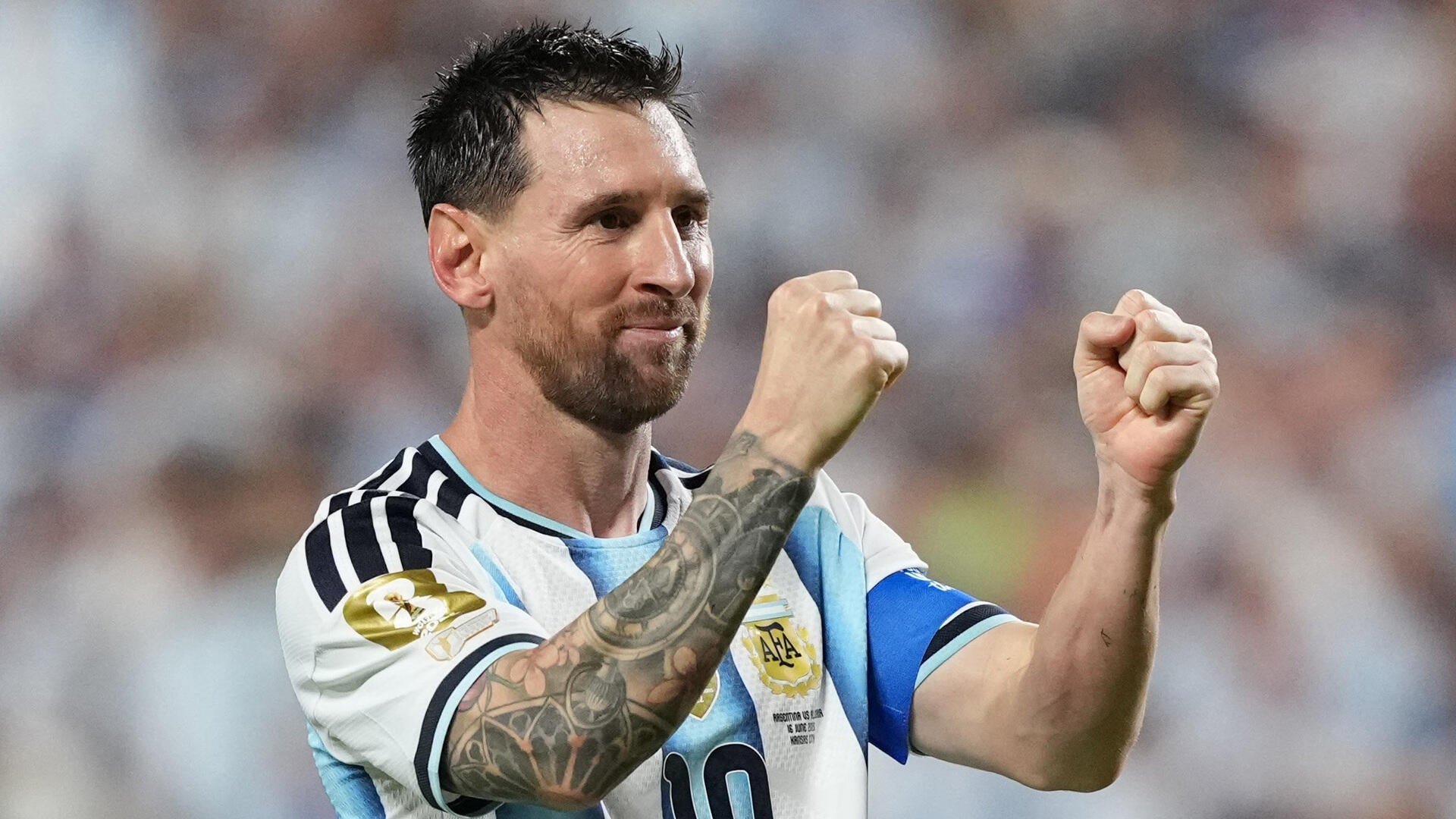 A bearded soccer player in a light blue and white striped Argentina jersey with "AFA" and a World Cup champions badge raises both fists in celebration before a blurred crowd.