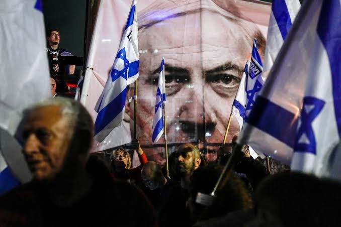 A crowd waves Israeli flags in front of a large banner displaying a close-up portrait of a man's face at an outdoor gathering.
