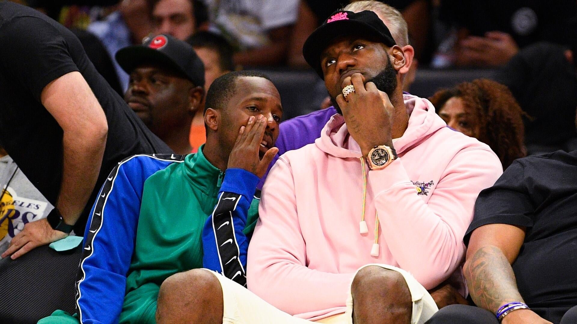 Two men sit courtside at a basketball game, one in a green and blue Nike tracksuit and the other in a pink hoodie and black cap, watching attentively among other spectators.