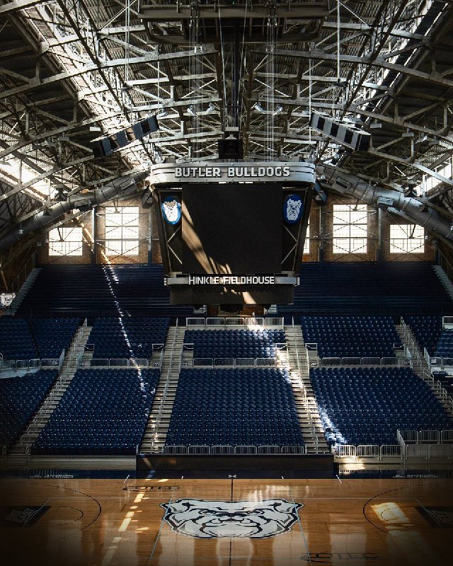 Empty basketball arena featuring a center-hung scoreboard labeled "BUTLER BULLDOGS" and "HINKLE FIELDHOUSE," with a bulldog logo at center court.