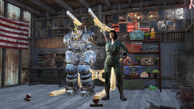 A video game screenshot shows a woman in a green jumpsuit standing beside a bulky blue power-armored figure inside a cluttered wooden workshop with an American flag on the wall.