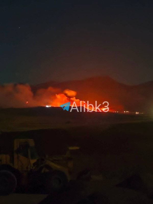 A distant nighttime fire burns brightly with orange flames and rising smoke behind dark mountainous terrain, while a piece of heavy machinery sits in the shadowed foreground.