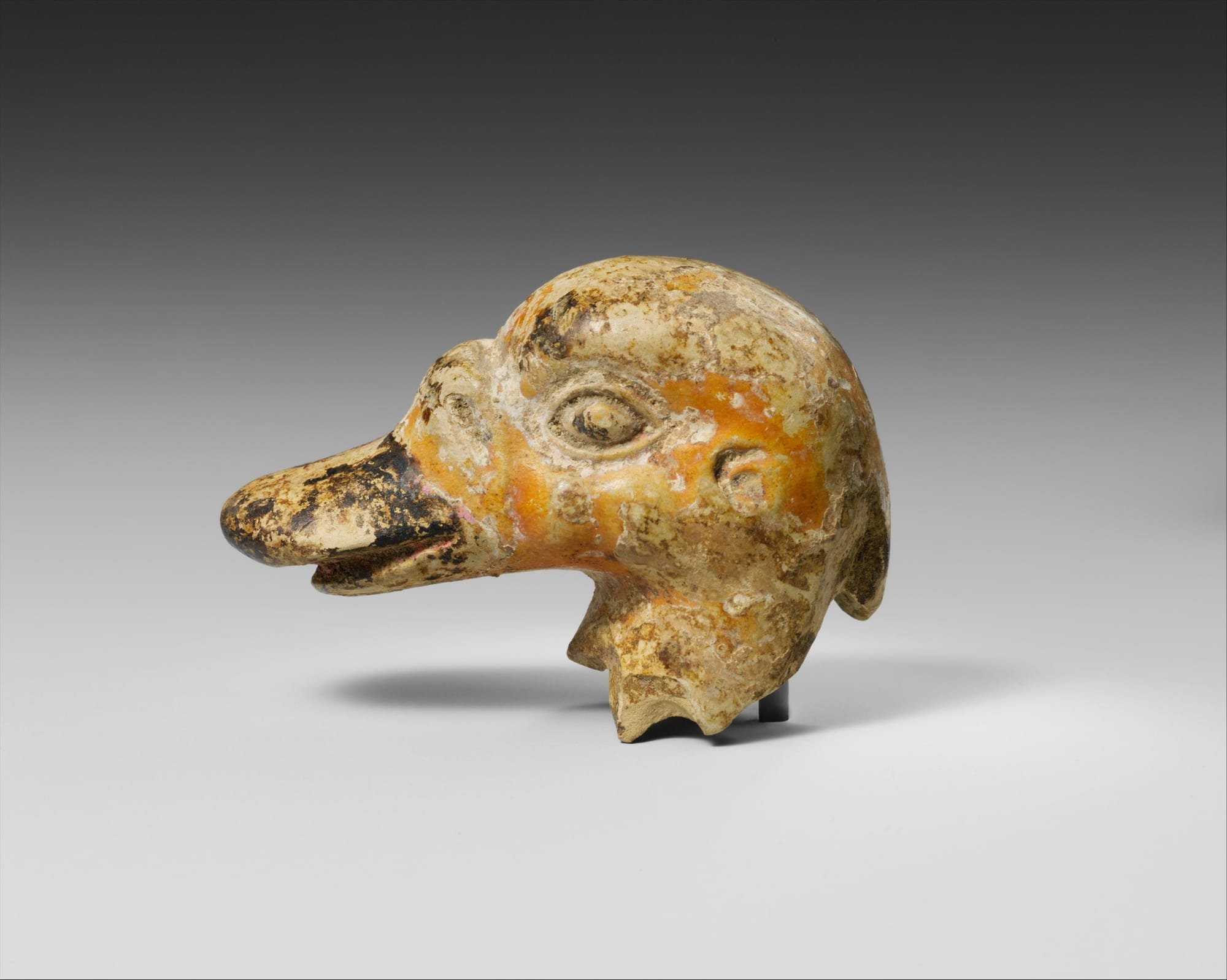 A weathered ceramic sculpture of a bird's head with an elongated beak, featuring orange and cream-colored glaze remnants, is displayed against a gray gradient background.