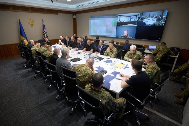 Military personnel and civilians in suits sit around a large conference table with documents, while a video feed displays on a wall screen near Ukrainian flags.