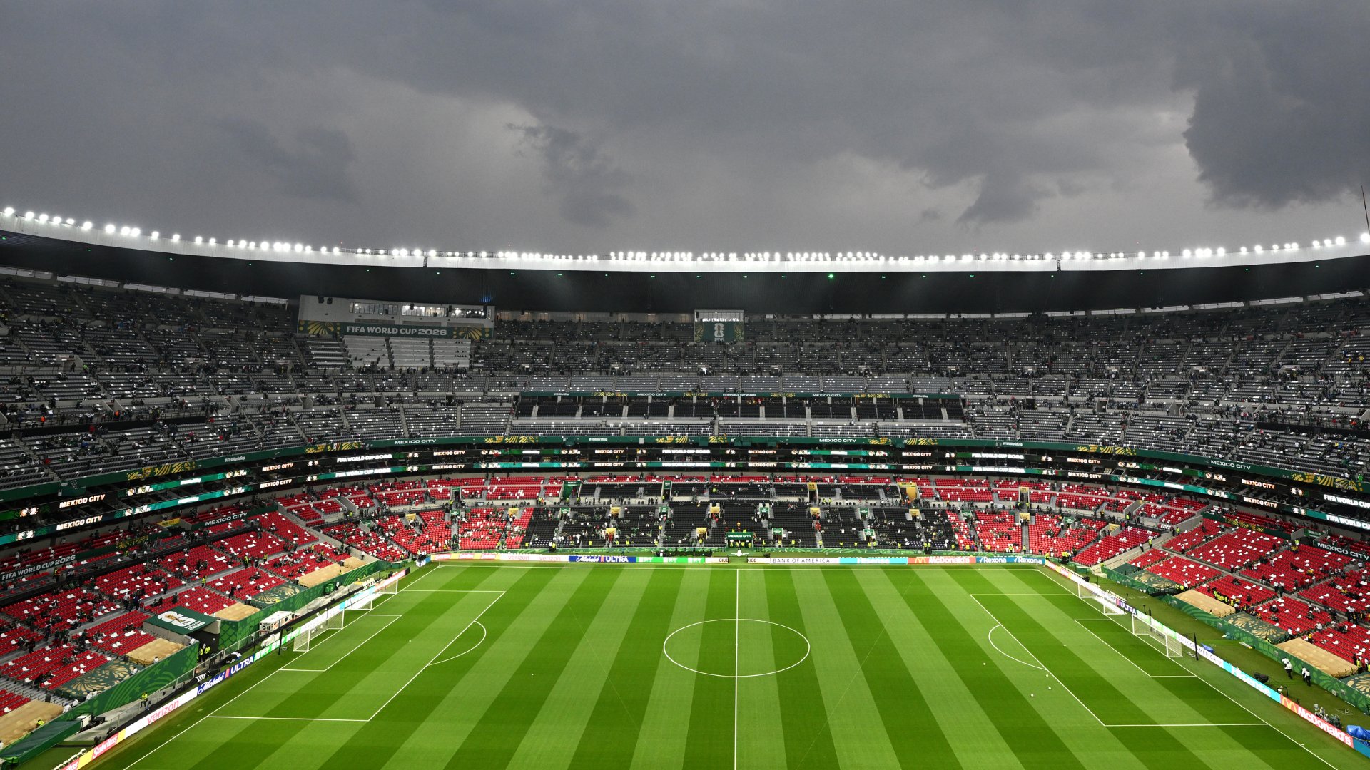 A nearly empty soccer stadium with red and gray seats surrounds a green pitch under dark, cloudy skies, with "FIFA World Cup 2026" and "Mexico City" signage visible.