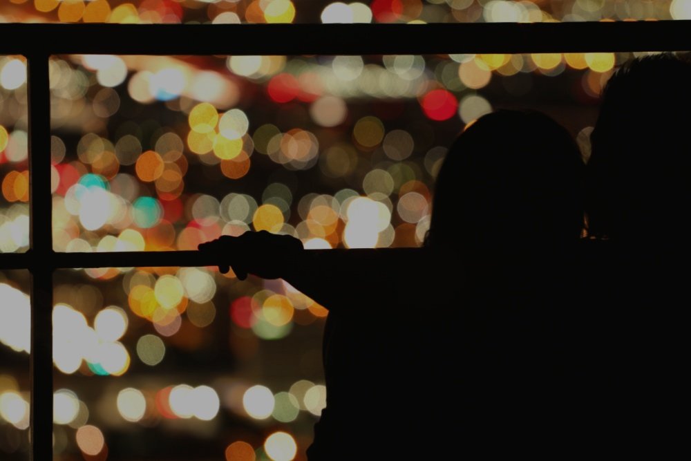 Silhouetted figures stand against a railing, framed by an out-of-focus backdrop of multicolored city lights at night.