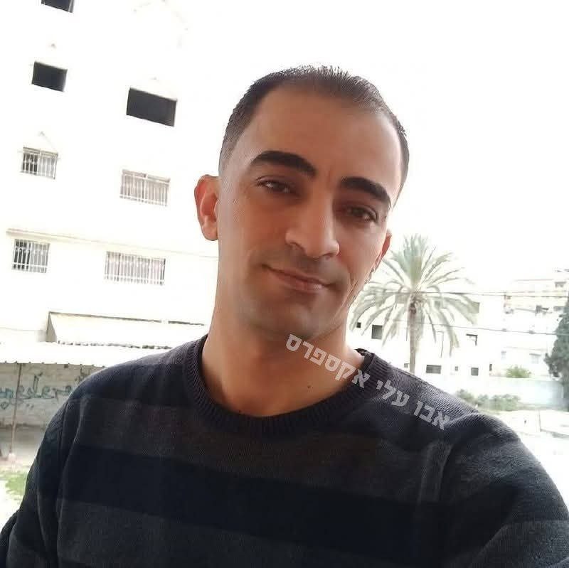 A man with short dark hair and a striped sweater poses for a close-up outdoor photo, with a light-colored building and a palm tree visible behind him.