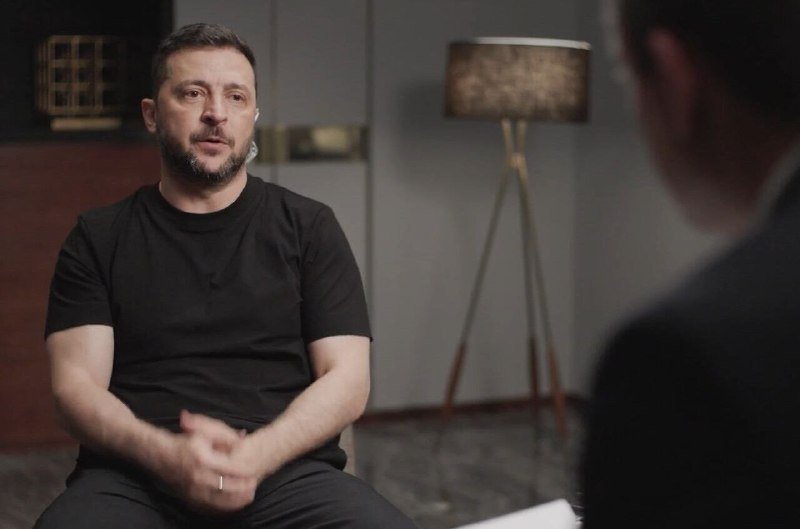 A bearded man in a black t-shirt sits indoors with an earpiece, speaking during an interview with a partially visible interviewer across from him.