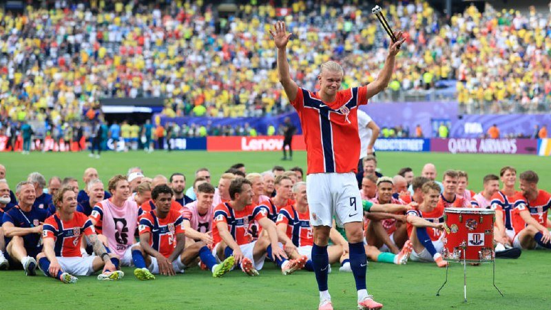 A player in a red and blue jersey (#9) raises both arms in celebration while teammates sit on the field, with a trophy nearby and a stadium crowd in the background.