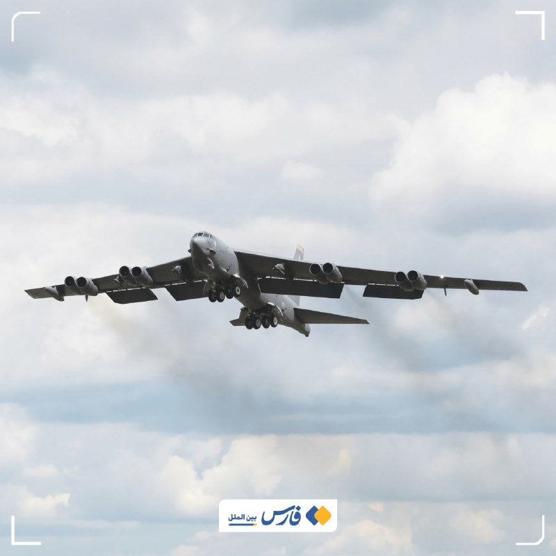 A large gray military bomber aircraft with eight engines flies through a partly cloudy sky, its landing gear extended.