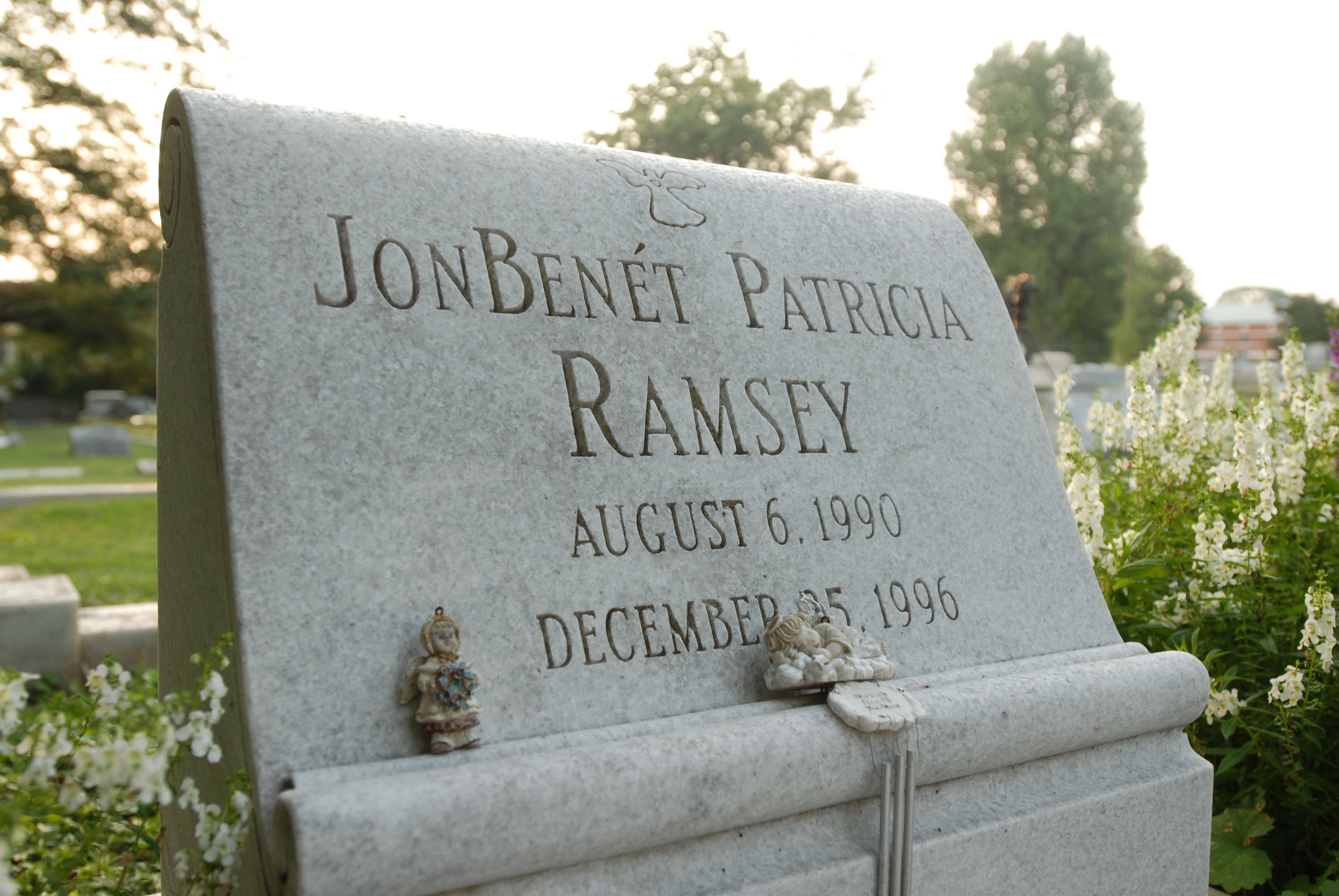 A granite headstone engraved with the name "JonBenét Patricia Ramsey" and the dates "August 6, 1990" and "December 25, 1996" sits in a cemetery, adorned with a small angel figurine and surrounded by white flowers and trees.
