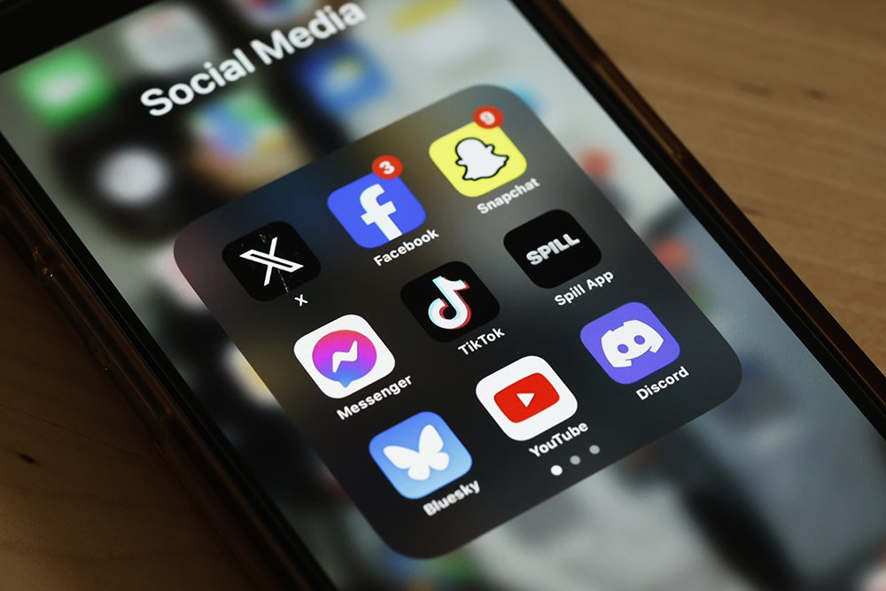 A smartphone displays an open "Social Media" folder containing app icons for X, Facebook, Messenger, TikTok, YouTube, Snapchat, Spill, Discord, and Bluesky.