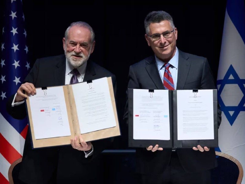 Two men in suits display signed documents and folders, standing between American and Israeli flags against a black backdrop.