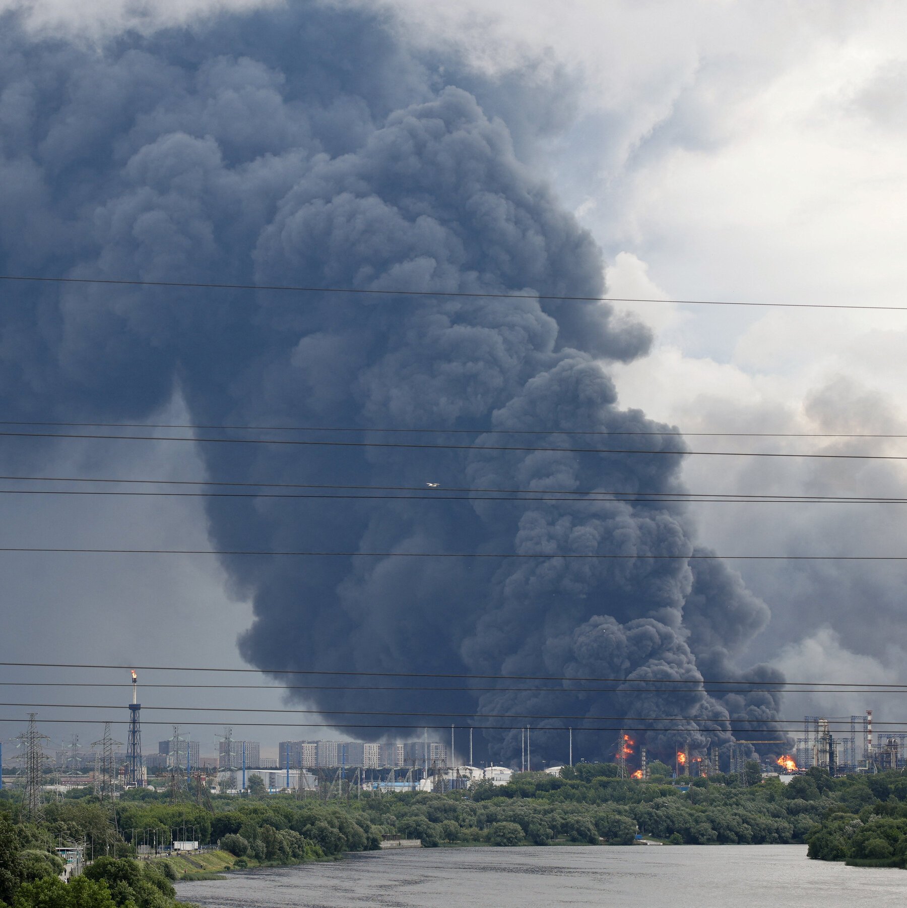 Thick black smoke billows from a fire at an industrial facility across a river, with visible orange flames, power lines, and an urban skyline in the background.