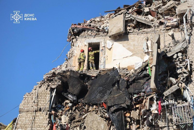 Two rescue workers in helmets and uniforms stand at the doorway of a heavily damaged multi-story brick building amid extensive rubble under a clear blue sky.