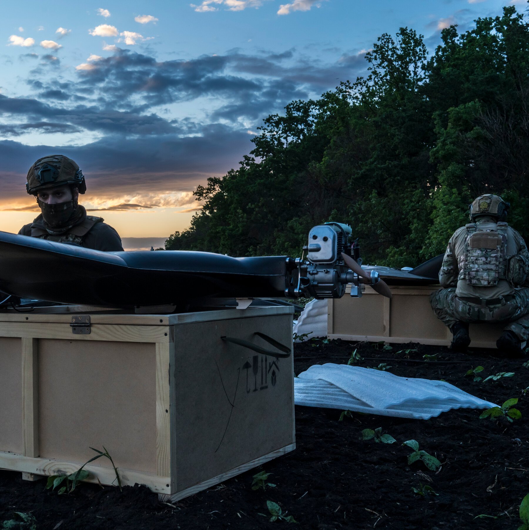 Two helmeted soldiers in camouflage gear unload a large drone from wooden shipping crates in a dirt field at dusk, with trees lining the background.