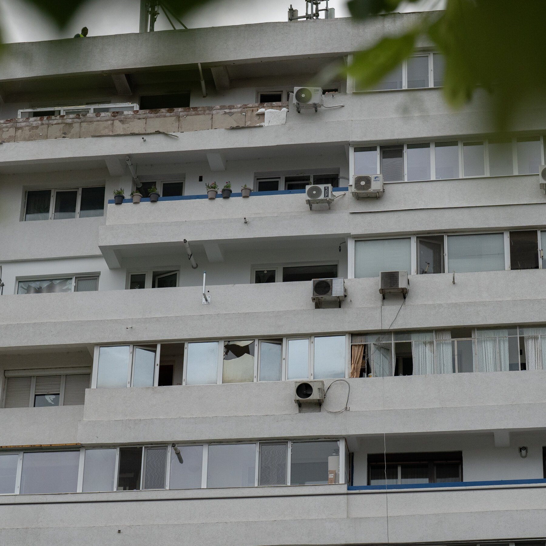 A multi-story white apartment building showing visible damage, including a broken window with shattered glass and exposed brickwork on an upper balcony, with multiple air conditioning units mounted on exterior walls.