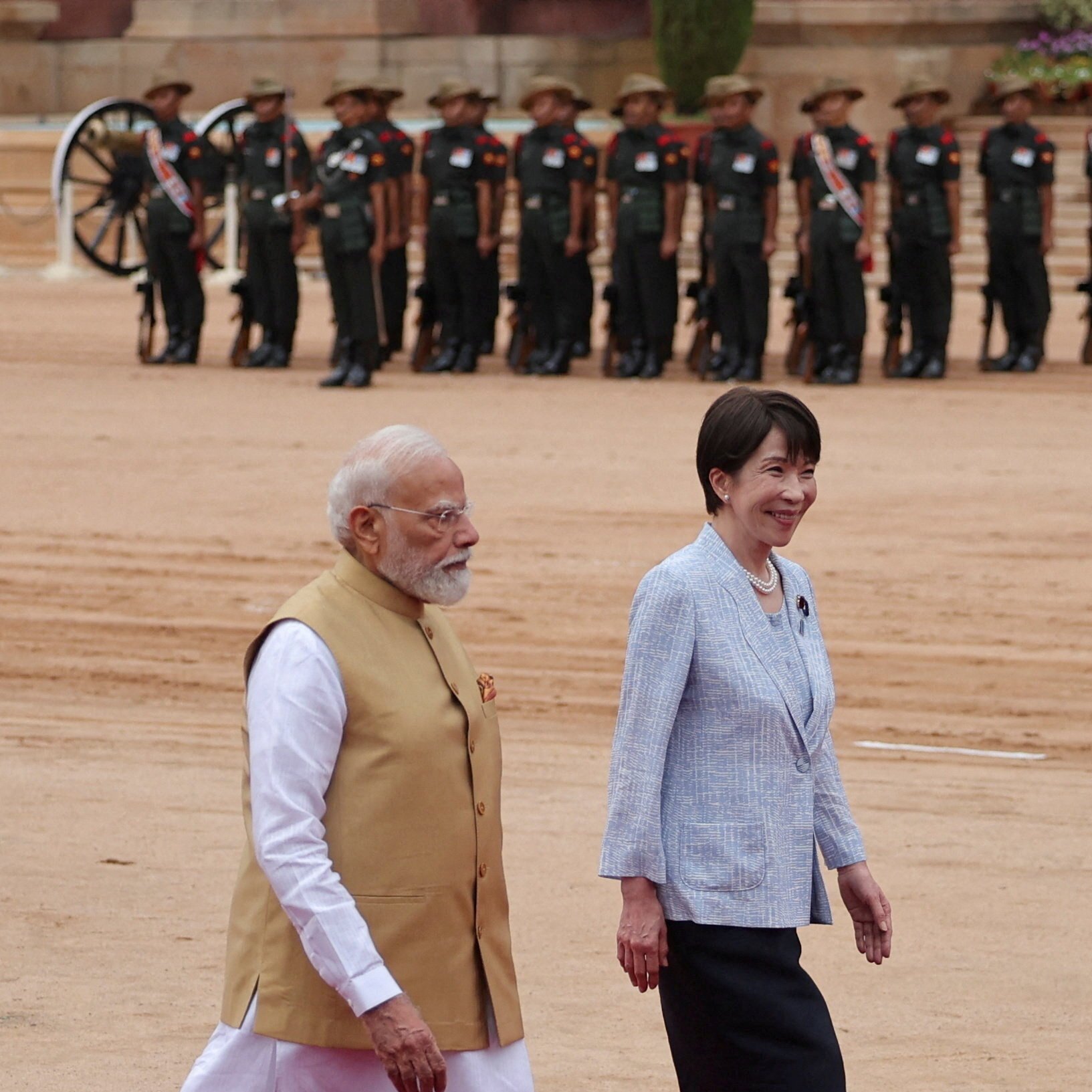 A bearded man in a tan vest and an East Asian woman in a light blue blazer walk side-by-side on a sandy parade ground, with rows of uniformed soldiers standing at attention behind them.