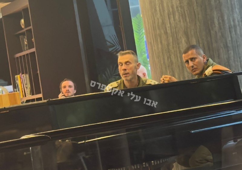 Three men in military-style shirts gather around a black grand piano in a wood-paneled room with bookshelves, viewed through glass bearing Hebrew text.