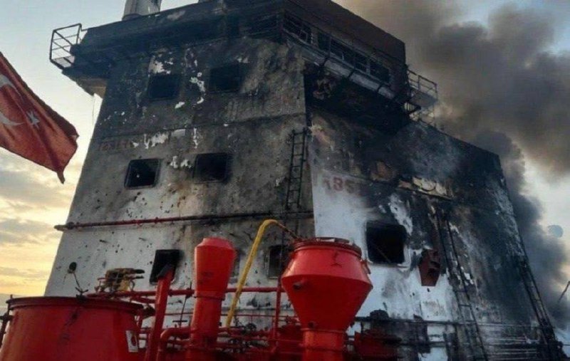 A fire-damaged, multi-story structure with blackened windows and billowing dark smoke stands beside a red industrial structure, with a Turkish flag visible on the left.