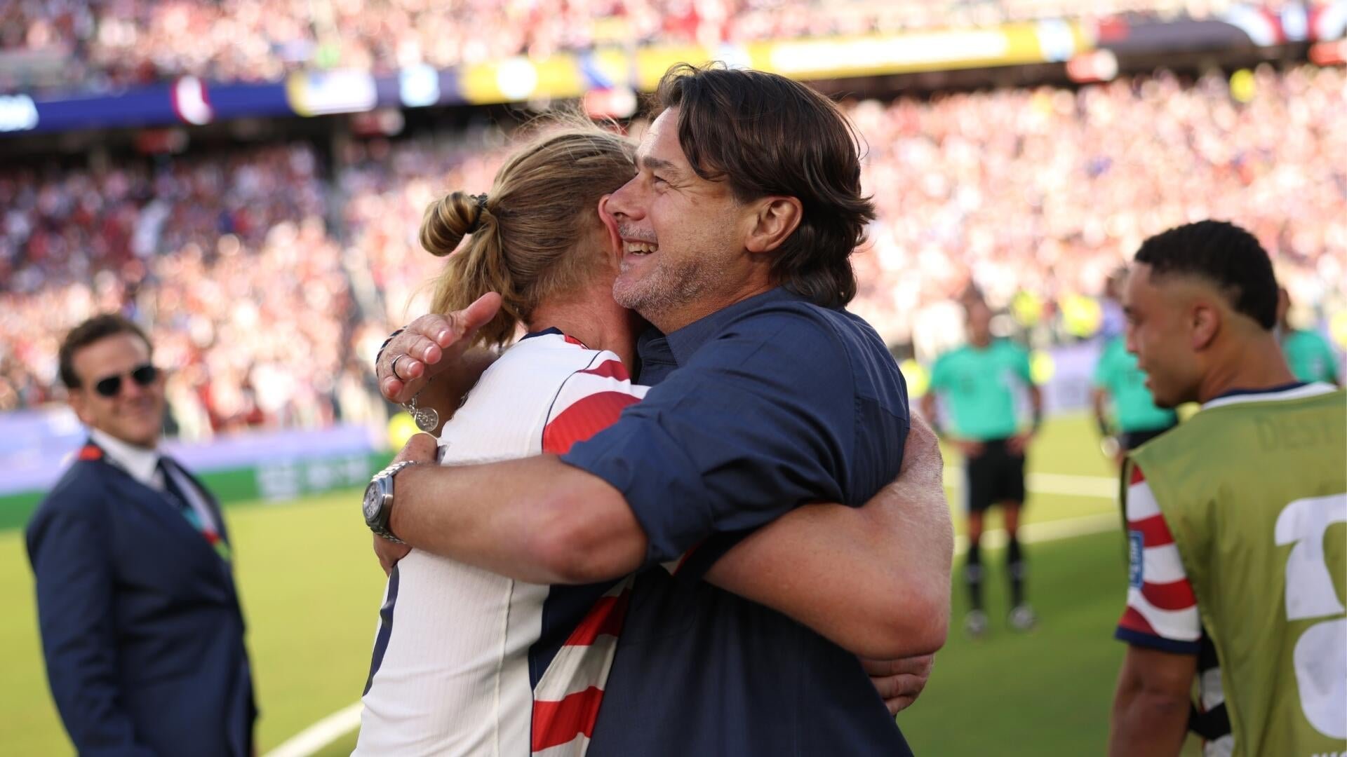 A man in a dark blue shirt hugs a player in a red, white, and blue striped jersey on a soccer pitch before a packed stadium crowd.
