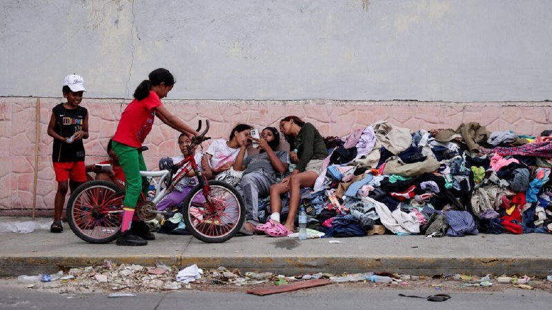 Children sit against a wall beside large piles of discarded clothing, while one girl holding a bicycle stands nearby on a littered street.