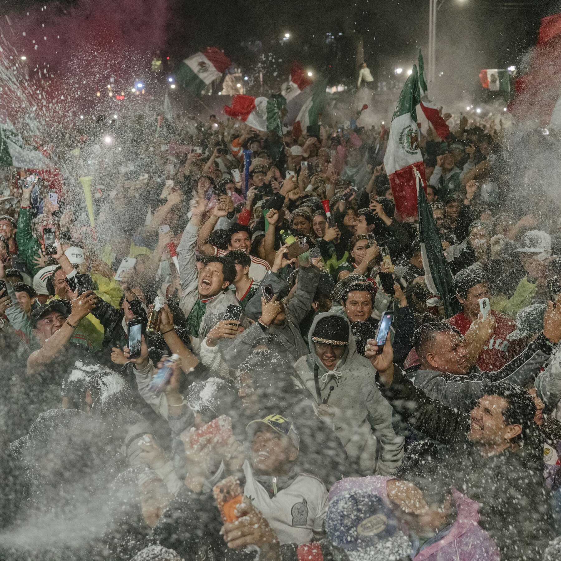 A dense crowd of cheering fans waves green, white, and red Mexican flags while spraying foam through a chain-link fence at night.