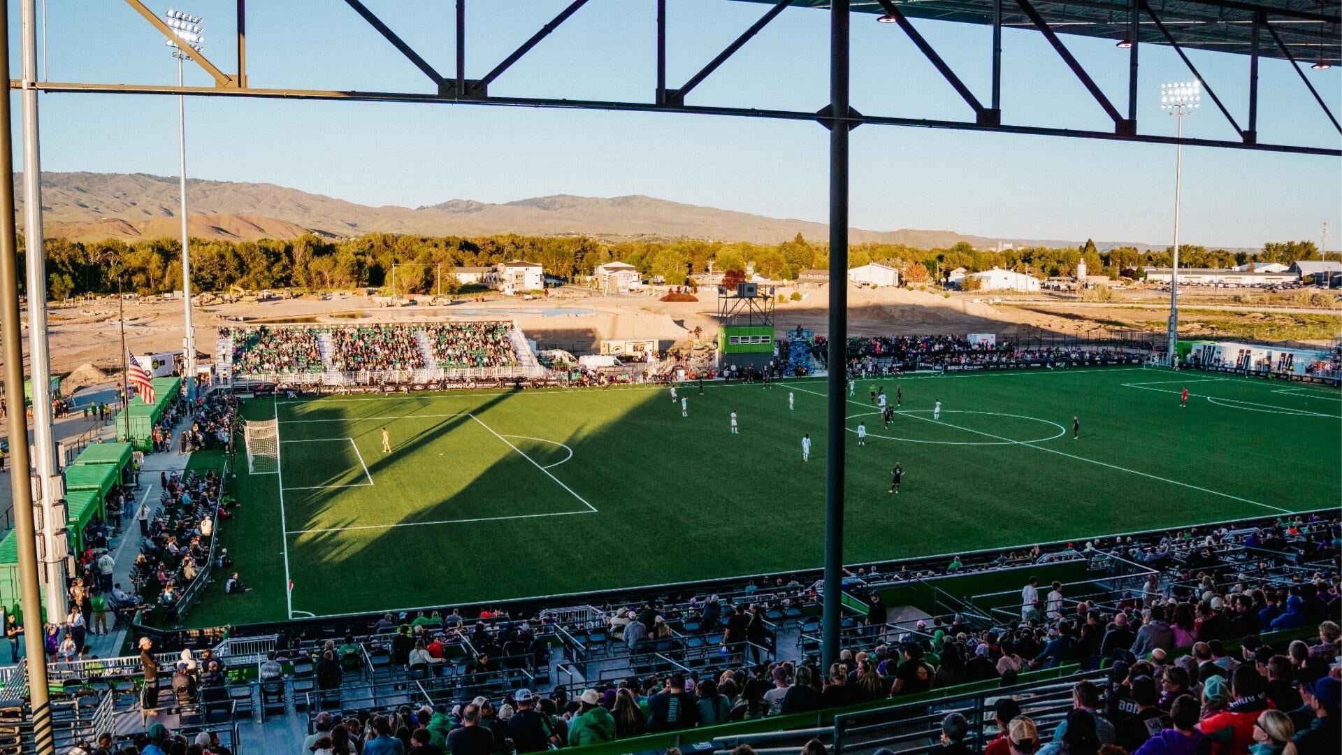 A soccer match unfolds on a green turf field surrounded by spectator-filled bleachers, with mountains and a clear blue sky visible in the background.