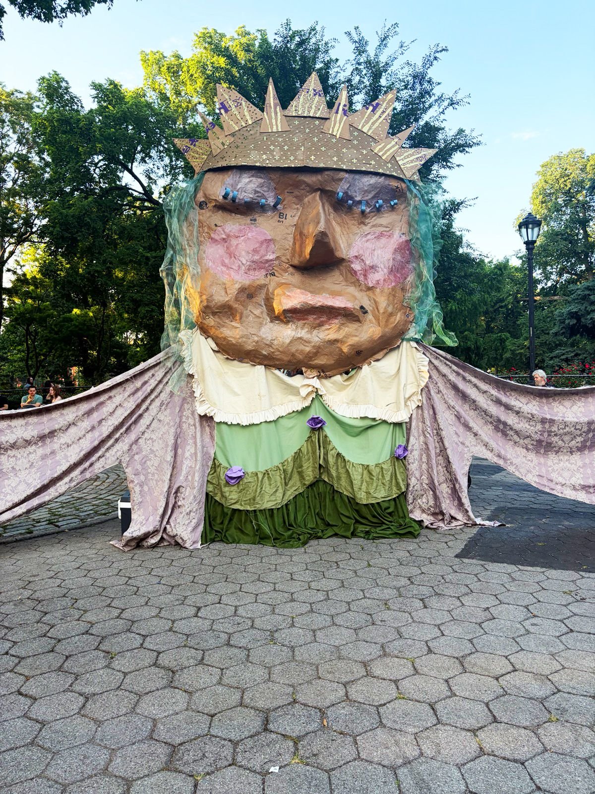 A large outdoor sculpture of a crowned figure with a papier-mâché face, green hair, and draped fabric base stands on a hexagonal-tiled plaza surrounded by trees.