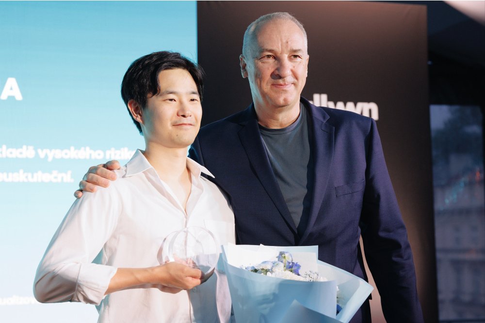 Two men pose together at an awards ceremony, one holding a glass trophy and a bouquet of flowers while the other has his arm around his shoulder, with a presentation screen behind them.