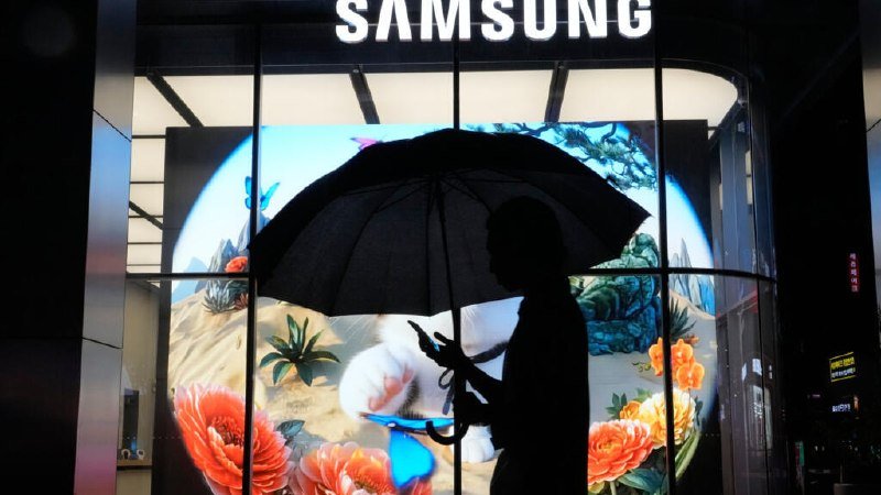 A silhouetted person holding an umbrella and a phone stands before an illuminated Samsung storefront display featuring a colorful floral scene.