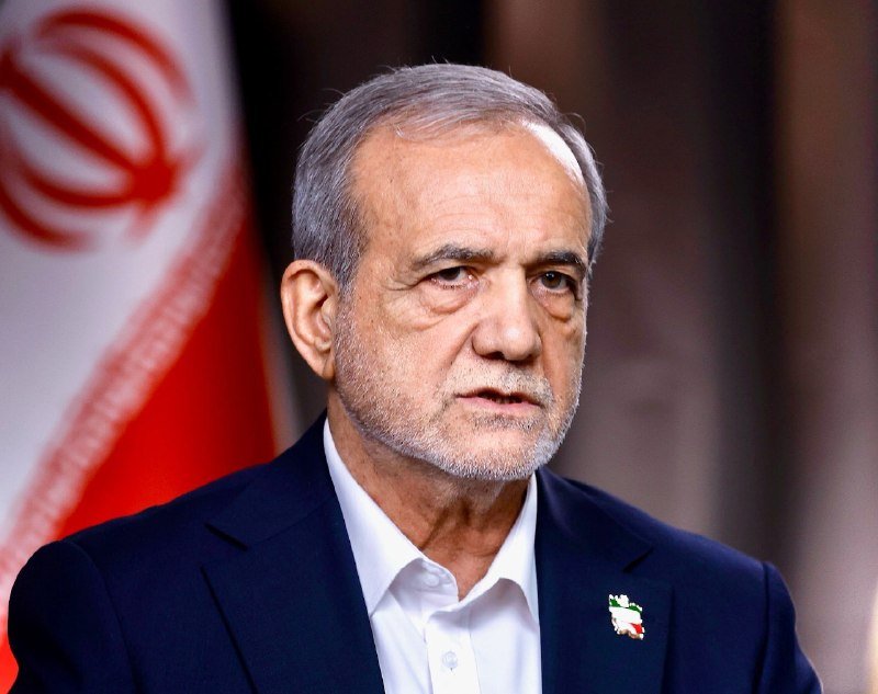 A gray-haired man in a dark suit and white shirt stands before an Iranian flag, with a small map-of-Iran pin on his lapel.