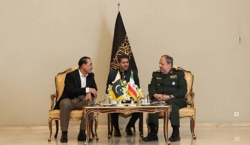 Three men, including one in military uniform, sit around an ornate table displaying Pakistani and Iranian flags with a black banner behind them.