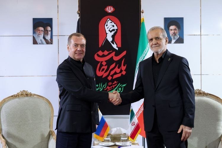 Two men in dark suits shake hands in front of the Iranian flag and a banner featuring a raised fist and Persian text, with Russian and Iranian flags on the table.