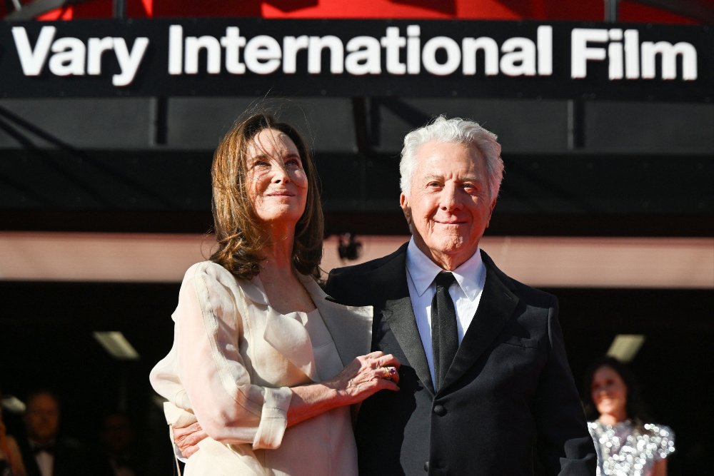 An older man in a black tuxedo and a woman in a white gown stand together beneath a sign reading "Vary International Film."