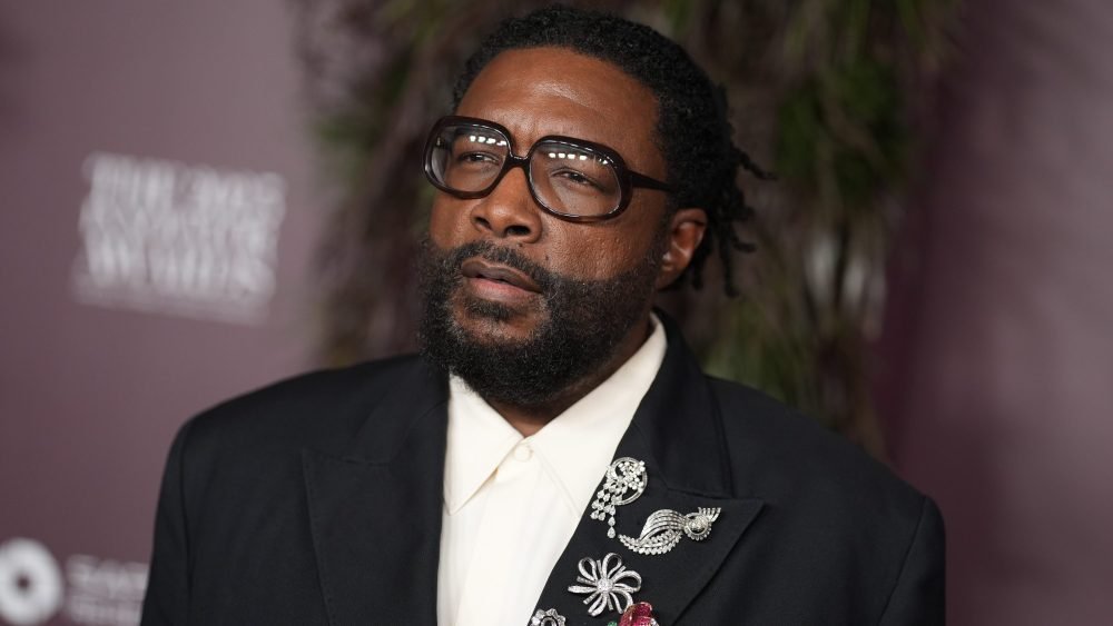A bearded man wearing black-rimmed glasses, a black suit jacket with jeweled brooches, and a white shirt poses against a maroon backdrop featuring partial white text.