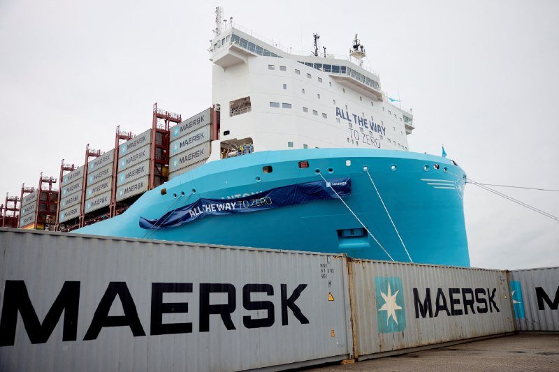 A large blue and white Maersk container ship displaying an "ALL THE WAY TO ZERO" banner is docked, with stacked Maersk shipping containers visible in the foreground.