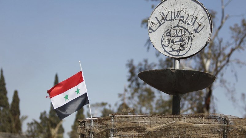 A Syrian flag flies beside a metal emblem with Arabic script, mounted on a structure surrounded by barbed wire and netting.