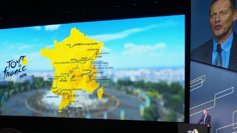 A man in a suit speaks at a podium beside a large screen displaying the "Tour de France 2026" route map overlaid on a map of France.