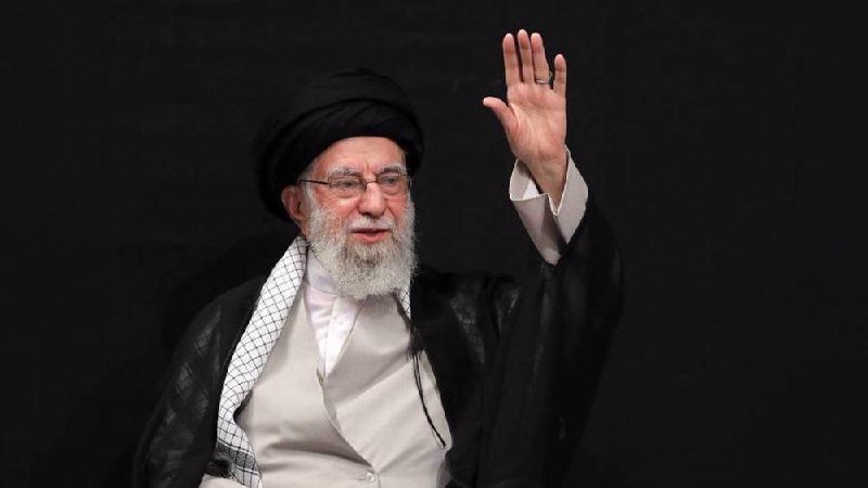 An elderly bearded man wearing glasses, a black turban, white robe, and dark shawl waves his raised hand against a dark background.