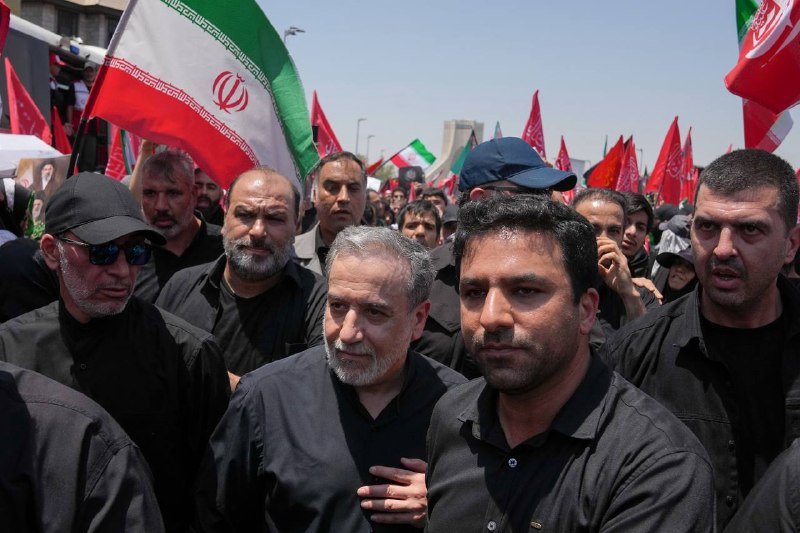 A crowd of men dressed in black gathers outdoors under a clear sky, waving Iranian flags and red banners in what appears to be a public procession.