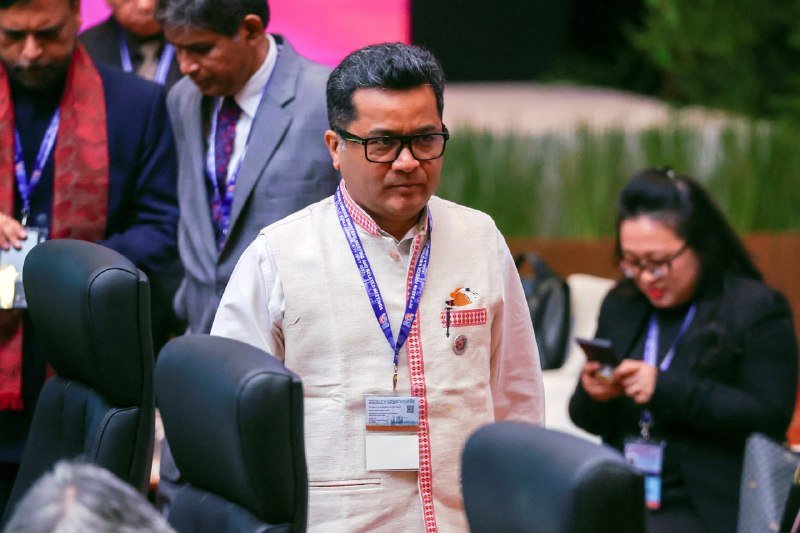 A bespectacled man in a cream vest and lanyard stands among seated attendees at a formal conference setting.