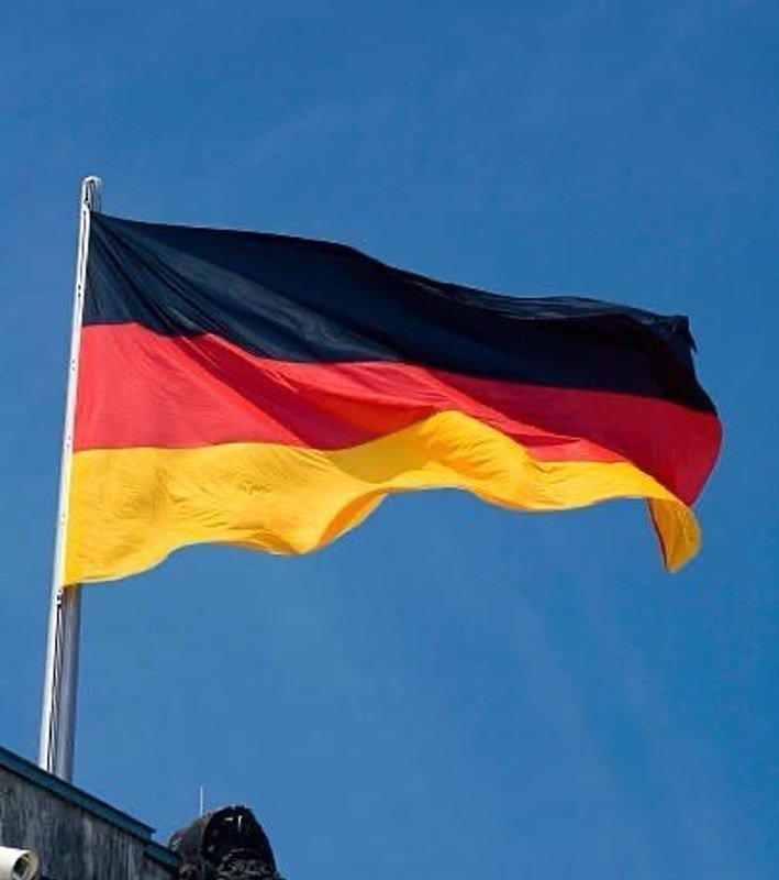 A German flag with black, red, and gold horizontal stripes waves on a flagpole against a clear blue sky, with a rooftop visible below.