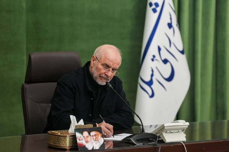A bearded, bespectacled man in black writes at a desk with a microphone, before an Iranian flag and green curtain.