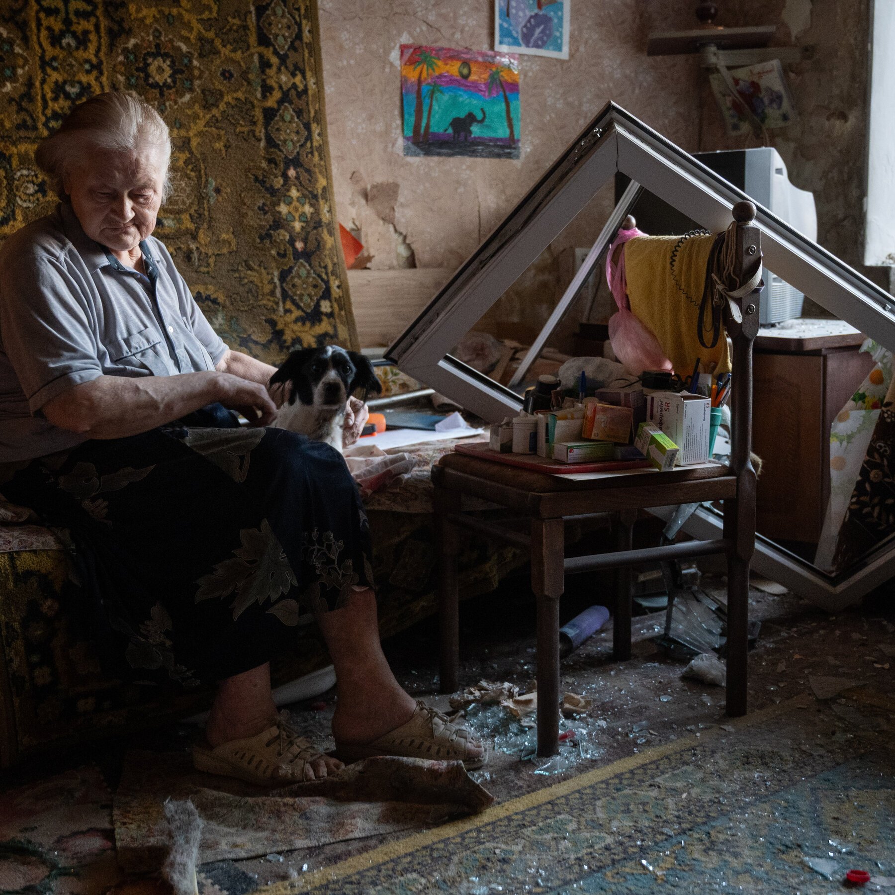 An elderly woman sits barefoot in a damaged room with shattered glass on the floor, holding a black and white dog beside a tipped-over table.