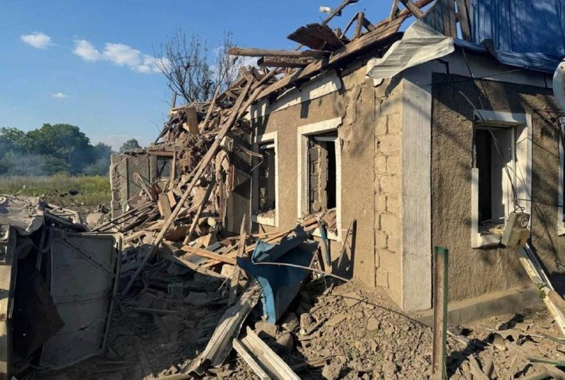 A heavily damaged single-story house sits amid scattered debris, with its roof collapsed, wooden beams exposed, and broken walls under a partly cloudy sky.
