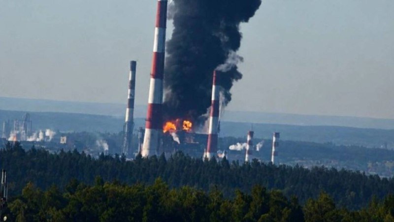 Black smoke billows from a fire at an industrial facility with tall red-and-white striped smokestacks, viewed beyond a forested foreground.