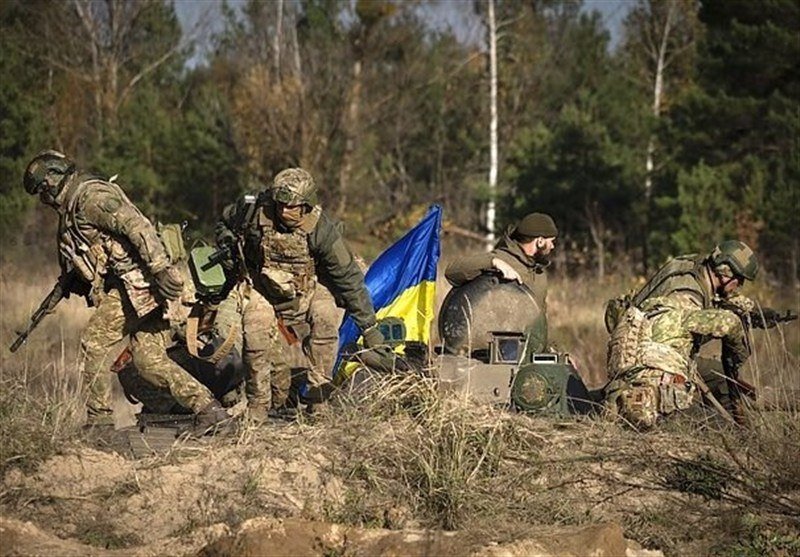 Soldiers in camouflage uniforms advance through a grassy field, taking cover behind a tank displaying a blue and yellow flag.
